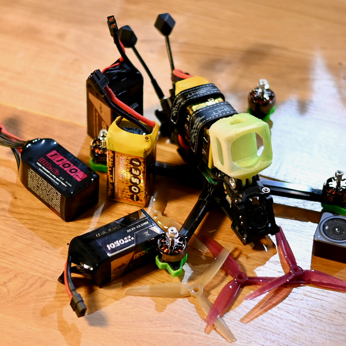 5 inch FPV freestyle quad on a workbench surrounded by LiPo batteries and spare propellers, showing battery leads and an action camera setup