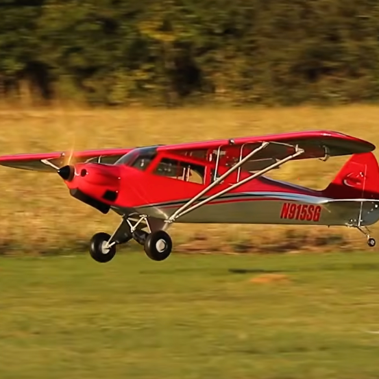Updated Hangar 9 Carbon Cub 15cc EP PNP 90-inch RC airplane in red and silver performing a low-altitude flight over a grass runway 复制